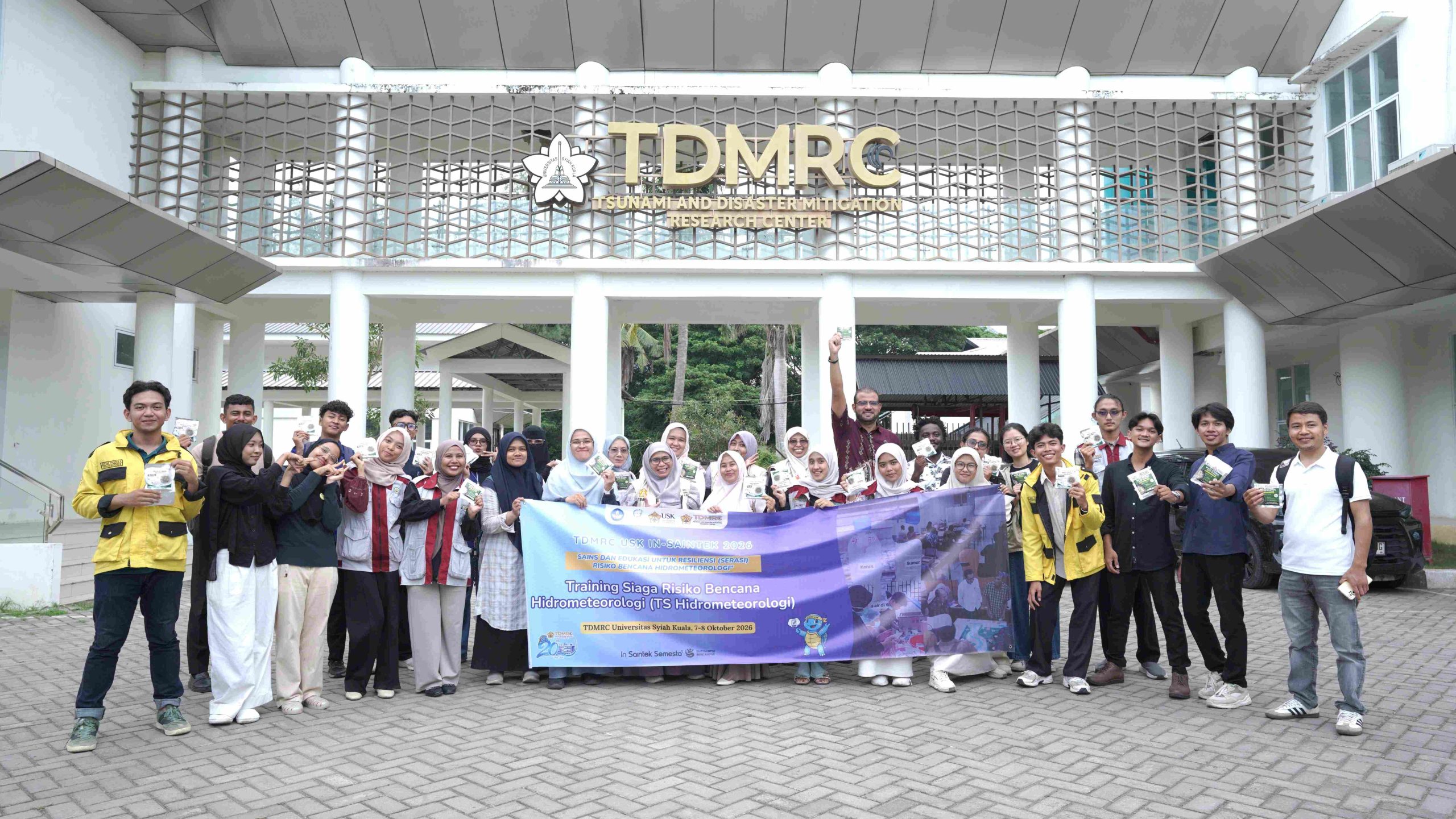 Students, international students, teachers, and the TDMRC USK team pose with the training banner and their packaged emergency food in front of the TDMRC USK building after the first day of training.